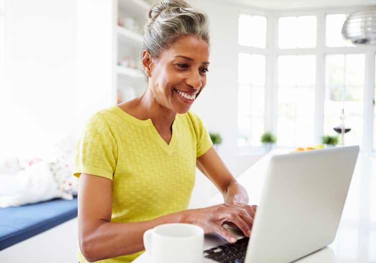 A grey-haired woman in a yellow shirt sits at a laptop with a white mug researching.
