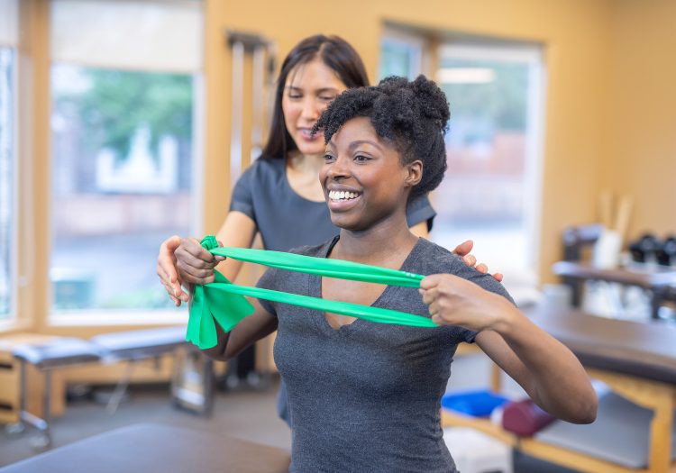 woman holding a resistance band with a female physical therapist behind her correcting her form