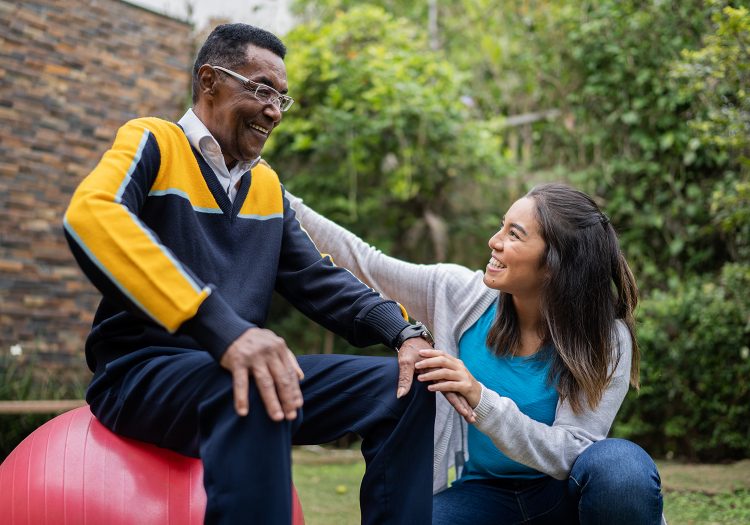 older man on an exercise ball with a woman helping him balance
