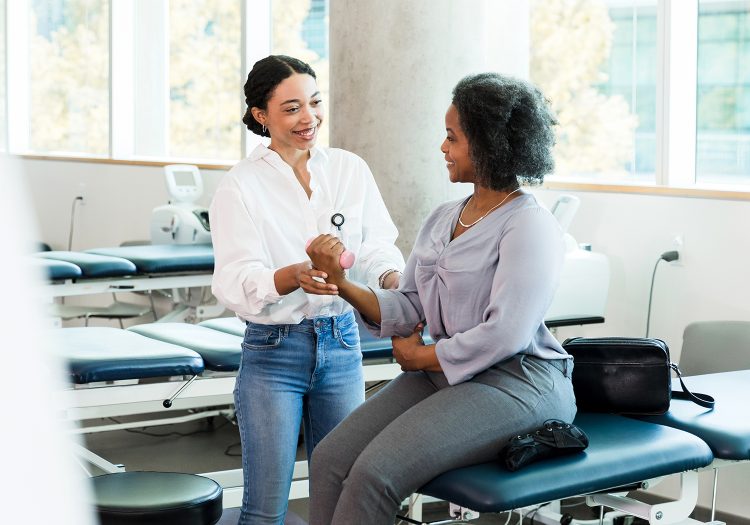 woman sitting on a physical therapy table holding a small hand weight, while female physical therapist guides her
