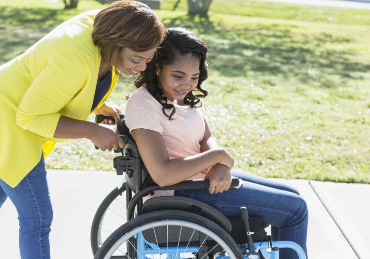 A woman assists a girl in a wheelchair through a park.