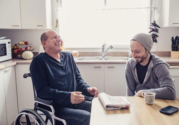 man in wheelchair laughing with a younger male in a kitchen at a table
