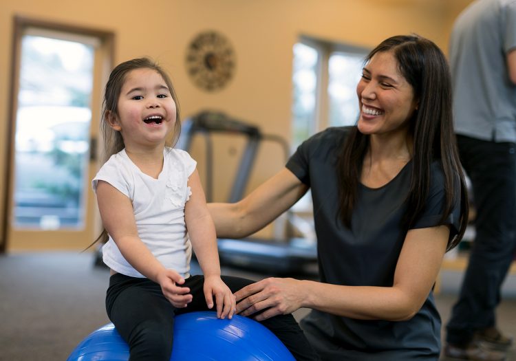 young girl sitting on medicine ball with a woman kneeling beside her helping her balance, both smiling