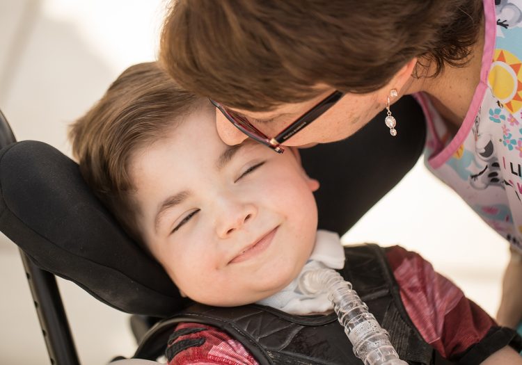 young boy in wheelchair smiling with a woman kissing him on the forehead