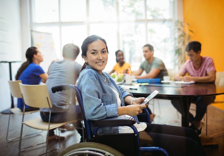 woman in wheelchair holding a phone smiling, people around a table in background